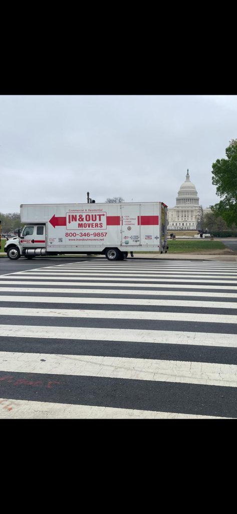 Side profile of an In & Out Movers truck driving across a wide pedestrian crosswalk in Washington DC, with the dome of the US Capitol building in the distant background.