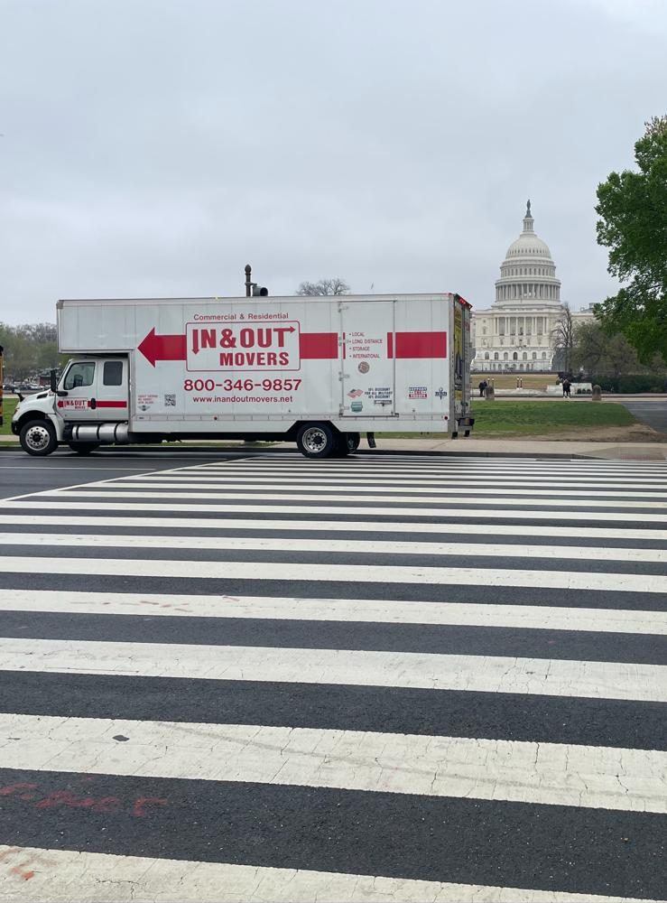 Side profile of an In & Out Movers truck driving across a wide pedestrian crosswalk in Washington DC, with the dome of the US Capitol building in the distant background.