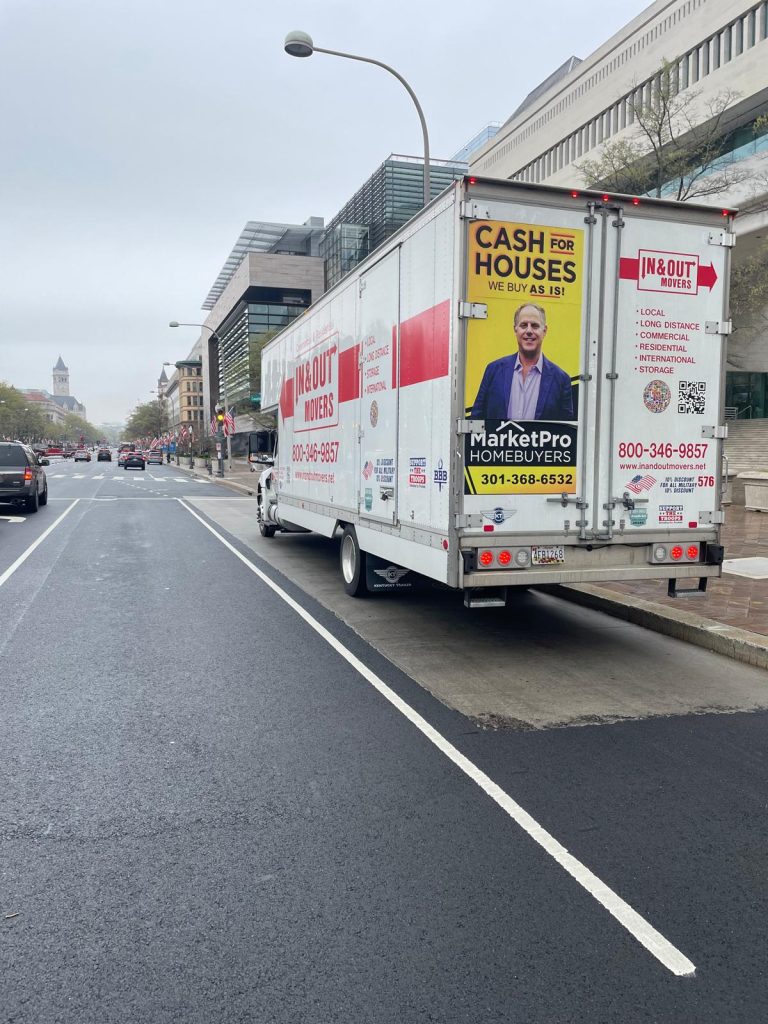 Rear view of a white In & Out Movers truck parked in Washington DC, featuring an advertisement for MarketPro Homebuyers and contact information for local and long-distance moving.