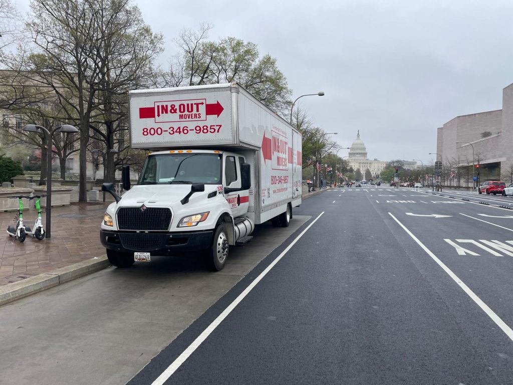 A white In & Out Movers truck parked on a street in Washington DC, with the iconic United States Capitol building clearly visible in the background under a cloudy sky.