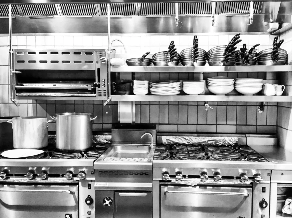 Commercial restaurant kitchen with stainless steel stoves, cookware, and shelves stacked with plates and pans.