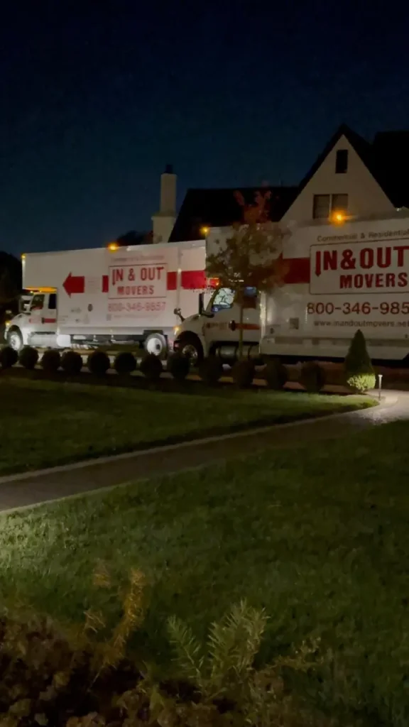 Two In & Out Movers trucks parked outside a residential home at night during a move-in or move-out service.