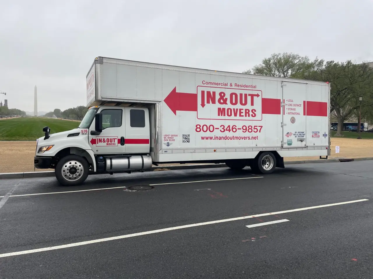 Side view of an IN & Out Movers commercial moving truck parked along a city street near a park.