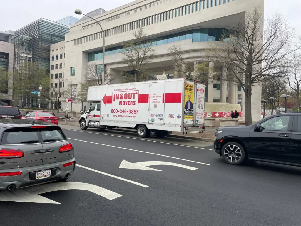 IN & Out Movers box truck parked on a city street near a large stone government building with passing traffic.