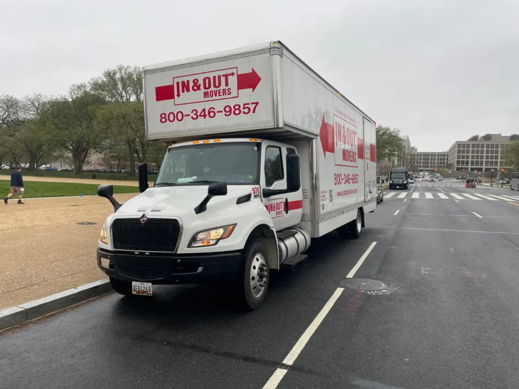 IN & Out Movers moving truck driving on a wide city roadway near grassy parkland on an overcast day.