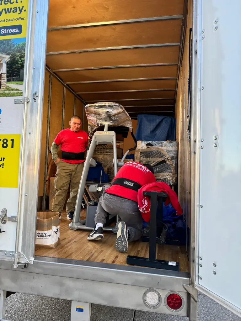 Two movers in red shirts loading wrapped furniture and equipment inside a moving truck