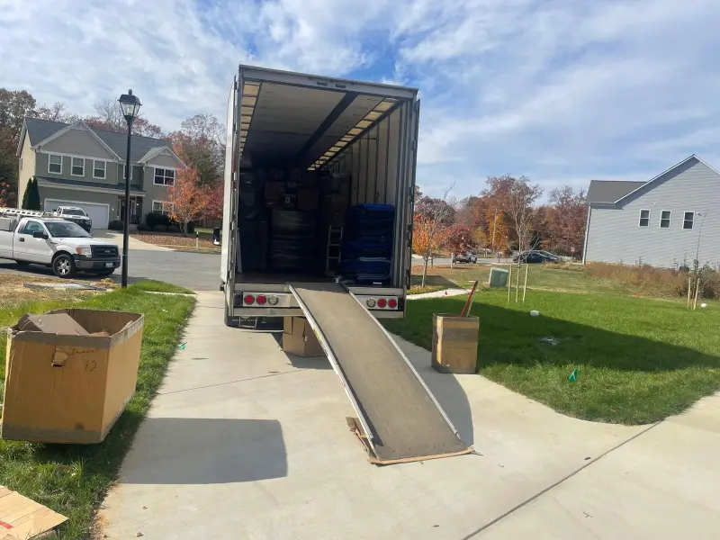 Open moving truck with a loading ramp extended onto a driveway, surrounded by cardboard boxes in a suburban neighborhood.