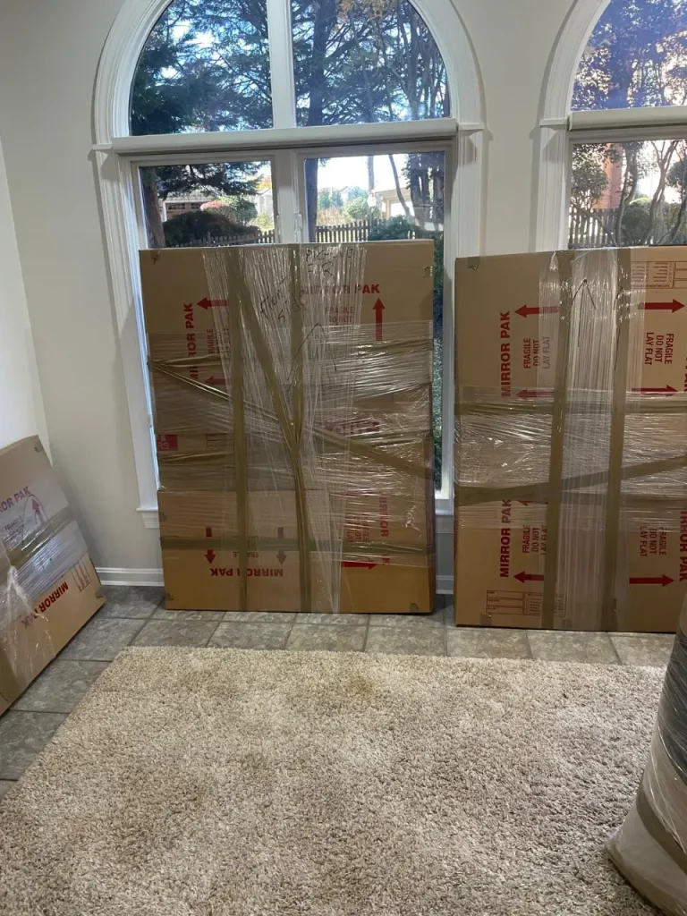 Stacked cardboard mirror boxes wrapped in plastic, prepared to pack for a move inside a home near large arched windows.