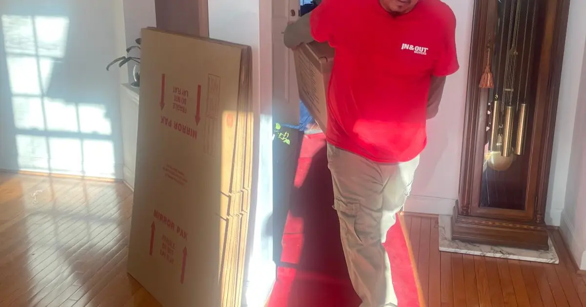 Mover carrying a packed box through a hallway beside stacked cartons during a home move with partial packing.