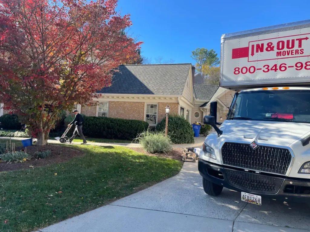 White glove movers unloading equipment from an In & Out Movers truck at a suburban home with autumn trees.